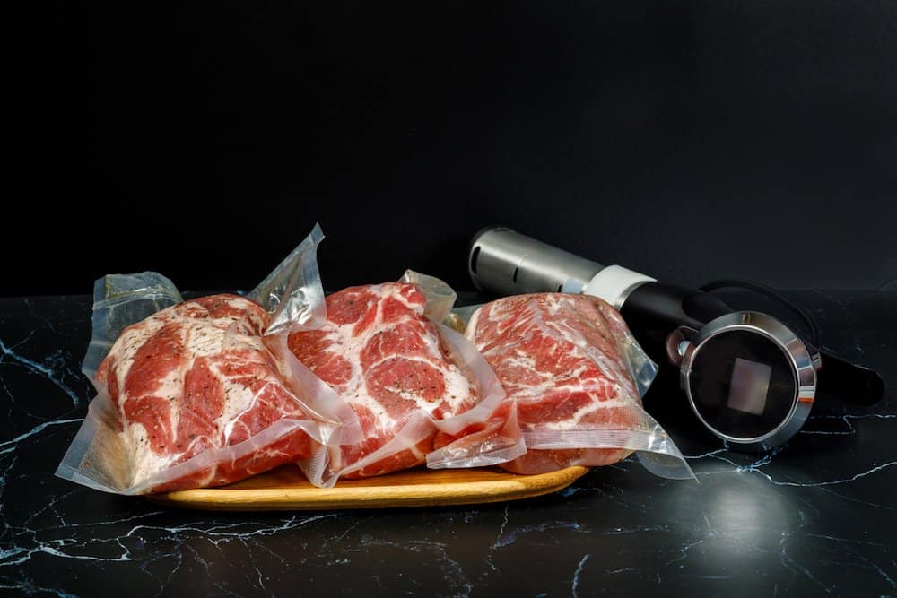 three steaks in sous vide bags on cutting board next to sous vide immersion circulator