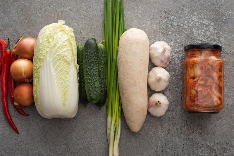 red chili peppers, onions, cabbage, cucumber, green onion, garlic, and jar of kimchi all in a row